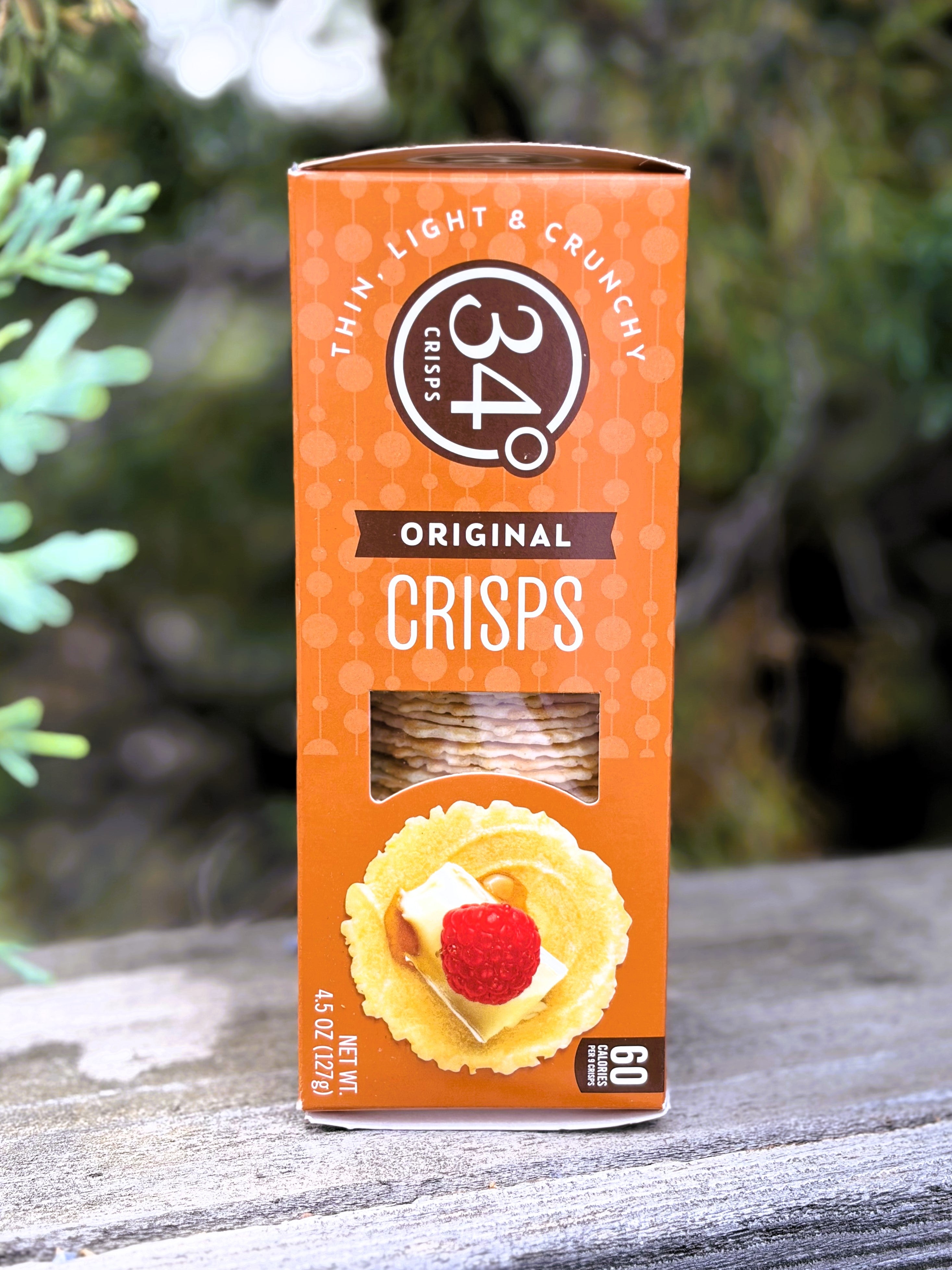 A box of 34° Original Crisps, Thin Light & Crunchy, standing upright on a weathered wooden surface with a blurred outdoor background of green foliage.