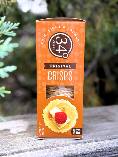 A box of 34° Original Crisps, Thin Light & Crunchy, standing upright on a weathered wooden surface with a blurred outdoor background of green foliage.