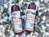 Two vacuum-sealed beef jerky sticks from Great Stone Ranch, one labeled 'Canyonstone Ranch Beefalo Salami' and the other 'Yellowstone Ranch Elk Summer Slices Beef Jerky', placed on a background of small gray pebbles.