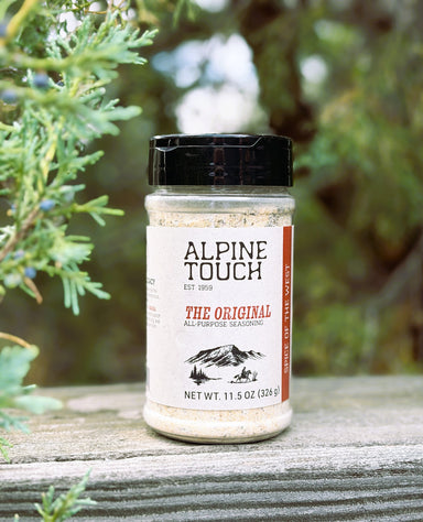 A bottle of Alpine Touch The Original All-Purpose Seasoning placed on a weathered wooden surface with green foliage in the background.