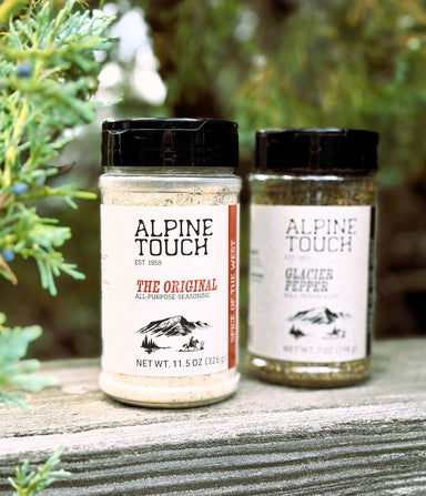 Two jars of Alpine Touch gourmet seasonings, 'The Original' and 'Glacier Pepper', displayed on a weathered wooden surface with green foliage in the background.