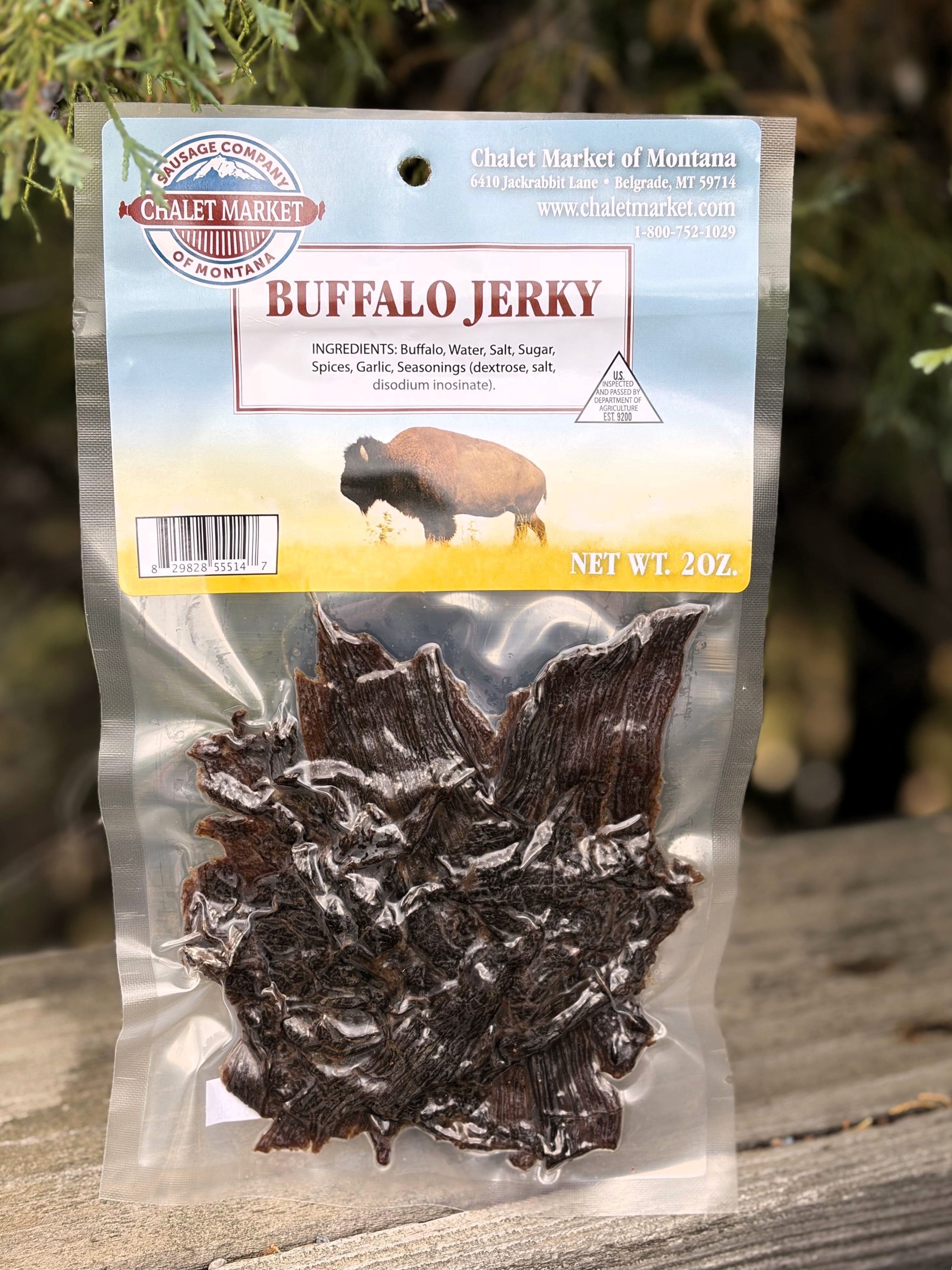 A 2 oz bag of Craft Market of Montana Buffalo Jerky, showing the dark brown jerky pieces inside a clear plastic pouch with a printed header card, set against a blurred outdoor background.