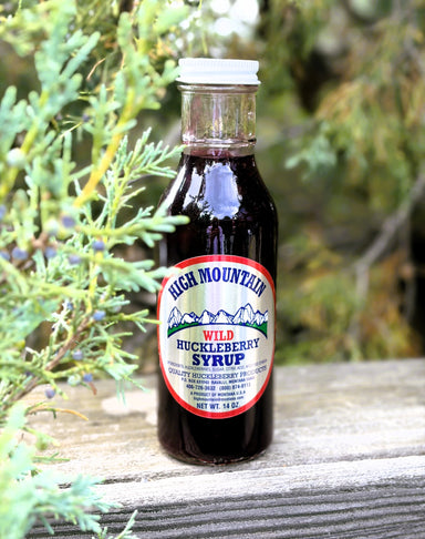 A bottle of High Mountain Wild Huckleberry Syrup standing on a weathered wooden surface with green foliage in the foreground and a blurred outdoor background.