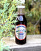 A bottle of High Mountain Wild Huckleberry Syrup standing on a weathered wooden surface with green foliage in the foreground and a blurred outdoor background.